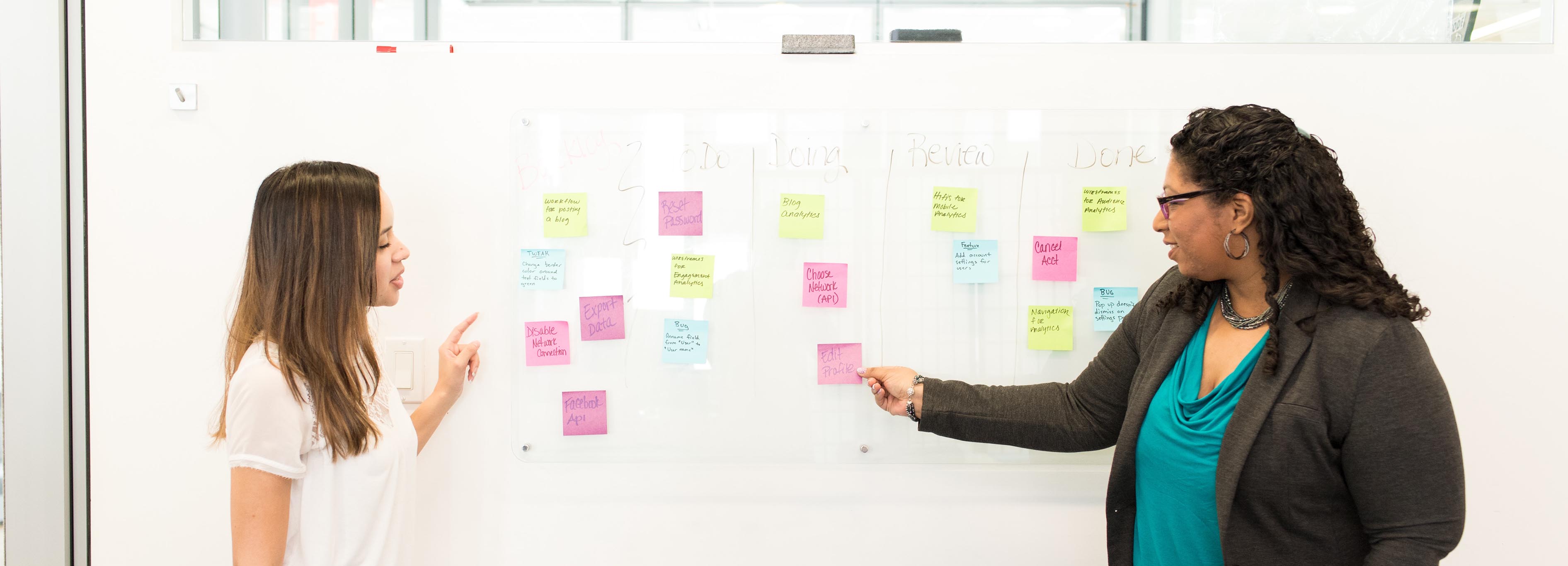 two female-presenting people gesturing at a whiteboard with stickies